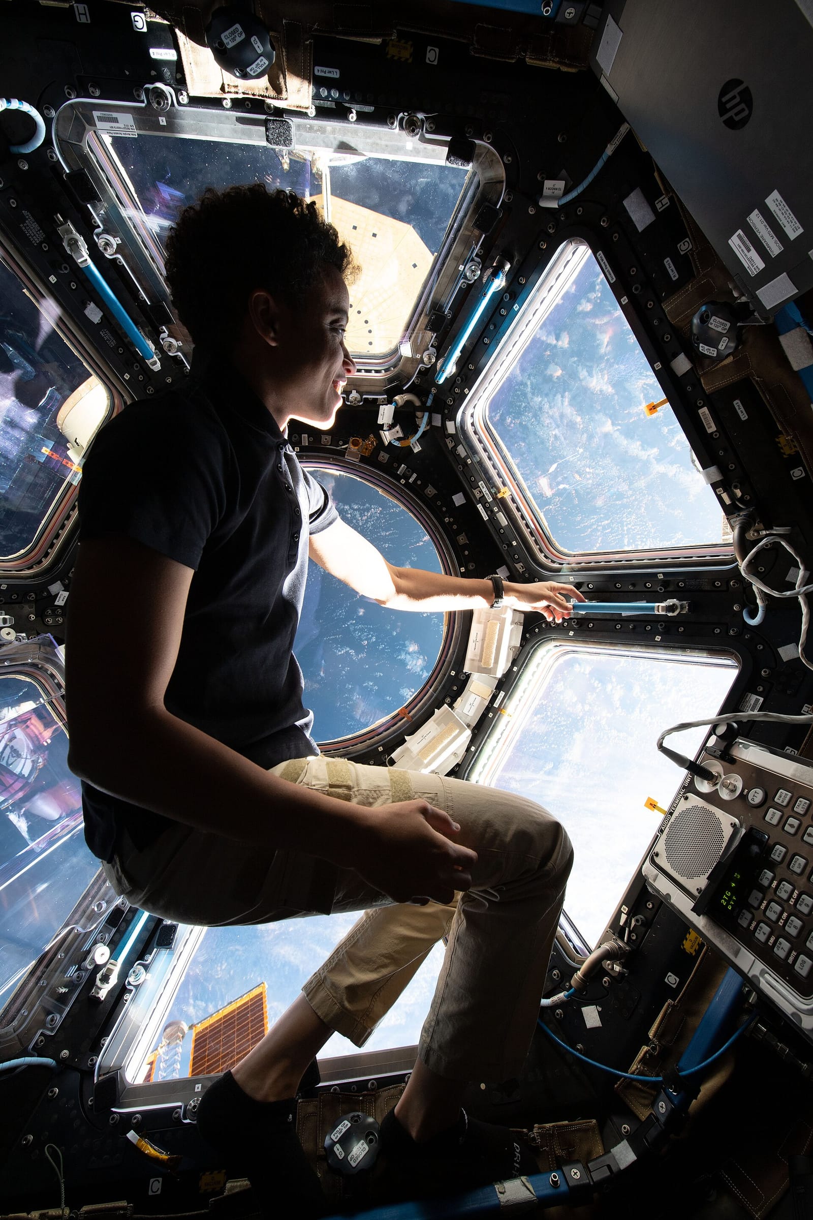 Astronaute en tenue sombre regardant par un hublot de la coupole, la Terre visible au-dessous.