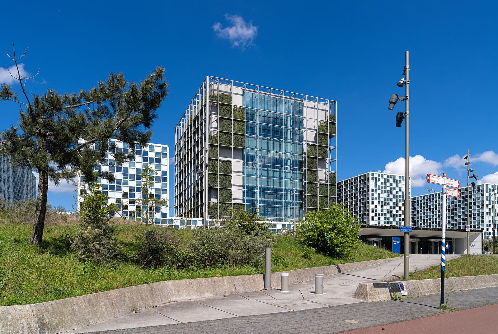 Immeuble vitré à façade végétalisée et bâtiments à carreaux bleus sous un ciel bleu, avec des arbres au premier plan.