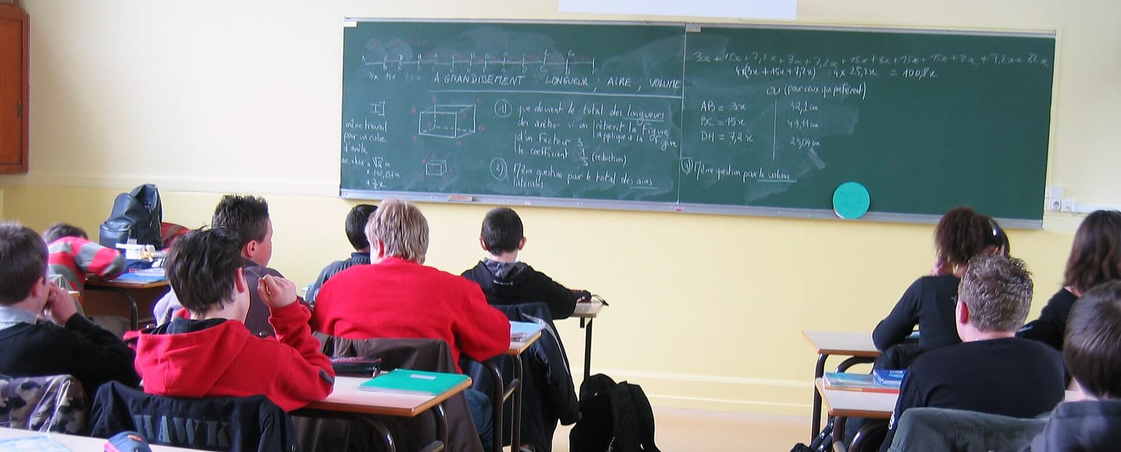 Élèves assis de dos dans une salle de classe, face à un tableau couvert de formules et de schémas.