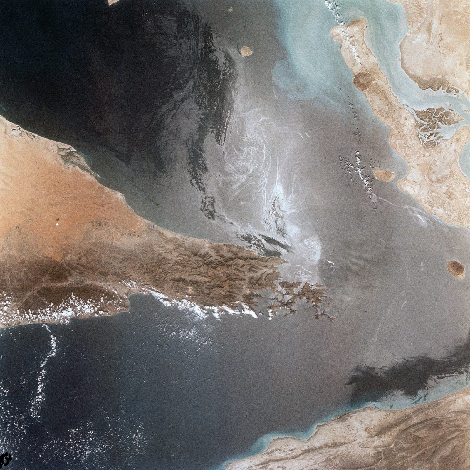 Vue aérienne du détroit d'Ormuz : côtes arides brun-ocre à gauche et en bas à droite, île et côte iranienne en haut à droite, mer grise et bleu sombre avec des 
