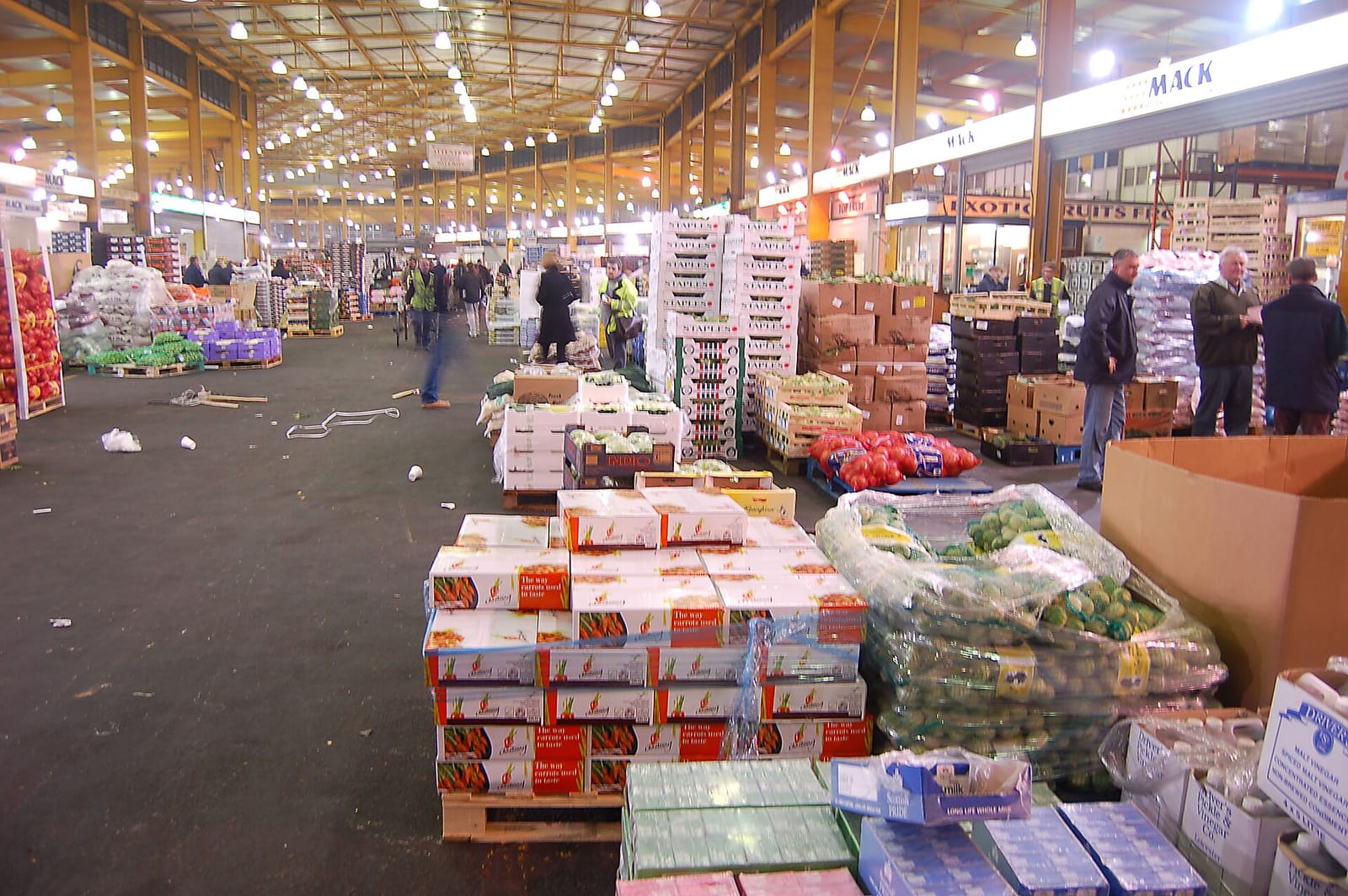 Halle d'un marché de gros éclairée, avec des palettes de caisses et de sacs de fruits et légumes au premier plan et des personnes au fond.