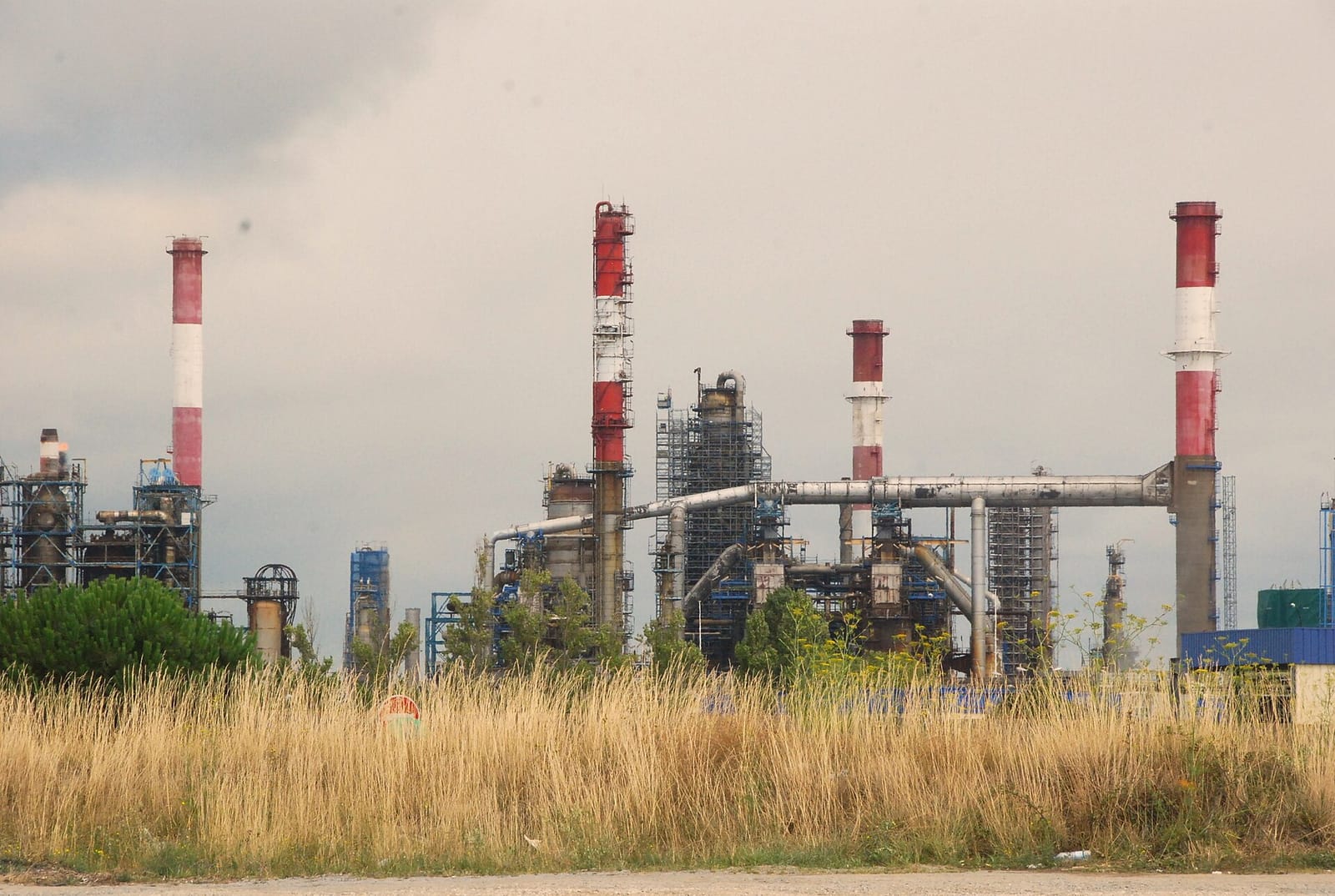 Installations d'une raffinerie avec des cheminées rouges et blanches, des échafaudages et des tuyauteries, derrière une prairie d'herbes hautes sous un ciel nua