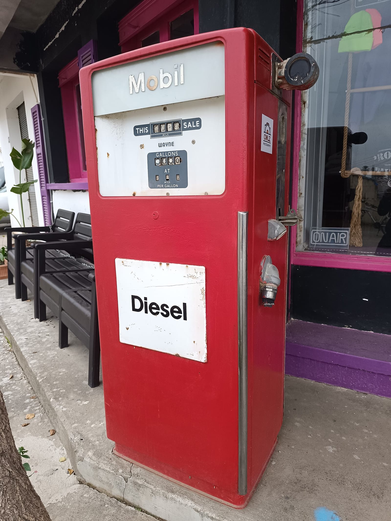 Ancienne pompe rouge Mobil avec une étiquette Diesel sur un panneau blanc, devant une vitrine.