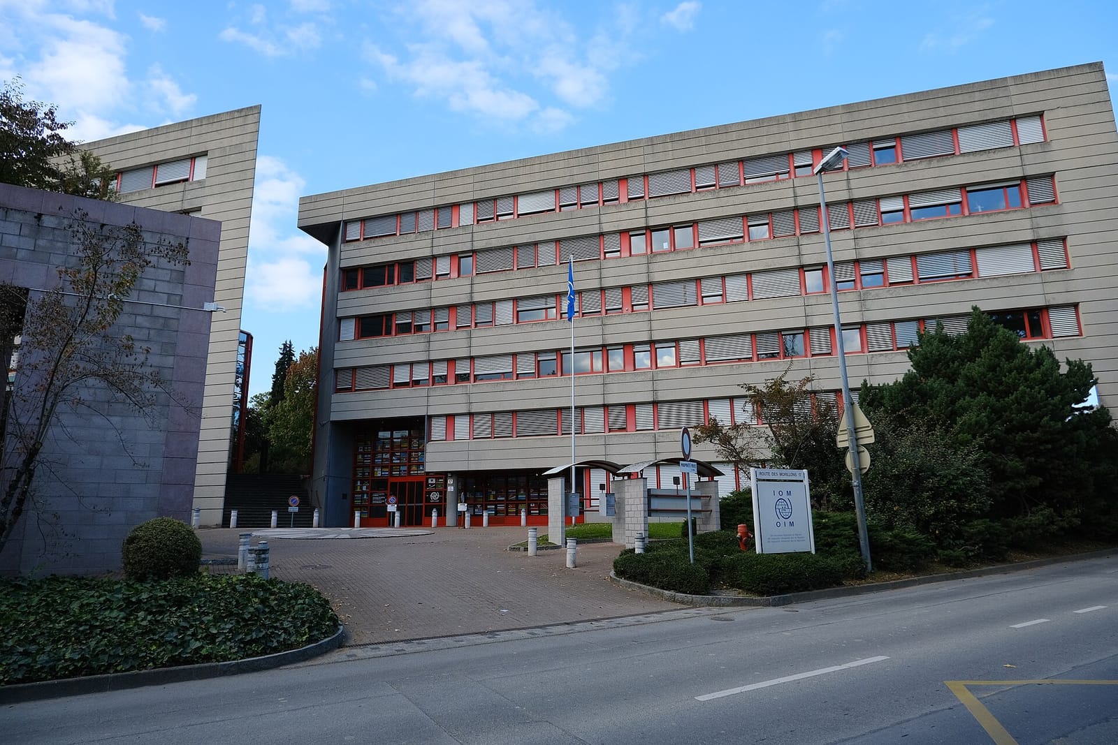 Façade d'un immeuble de plusieurs étages en béton clair, aux encadrements de fenêtres rouges, avec un mât portant un drapeau et un panneau « IOM OIM » devant l'
