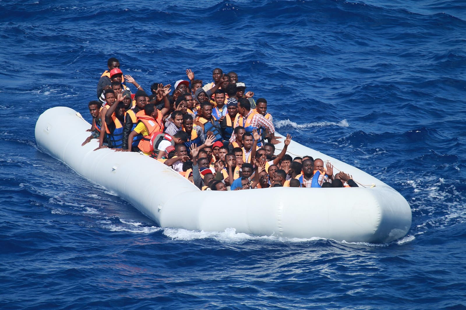 Canot pneumatique blanc surchargé de migrants en mer, dont plusieurs lèvent la main vers l'objectif.